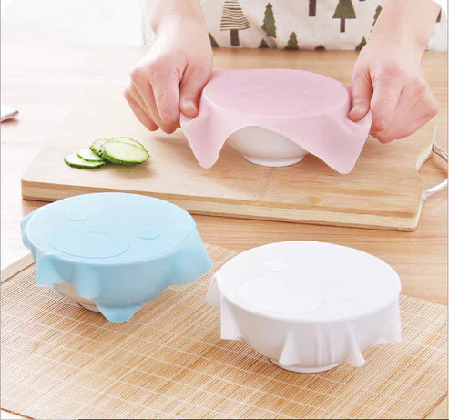 Three silicone food covers in different colors on a wooden surface.